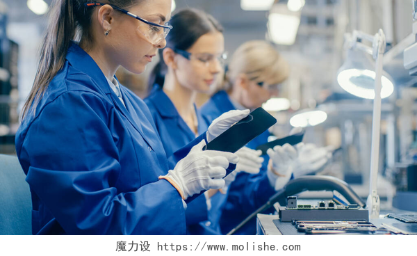 三个在工厂里工作的女人蓝色工作服和防护眼镜中的女性电子工厂工人采用智能手机屏风并进行质量检查。拥有多名员工的高科技工厂设施.